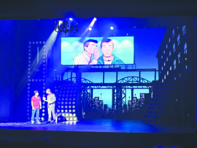 two actors on darkened stage with blue light, cityscape behind them, screen at top of stage showing two comic style characters talking