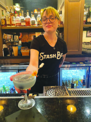 young female bartender with glasses and branded Stash Box t-shirt, standing behind bar, holding out martini glass with garnish