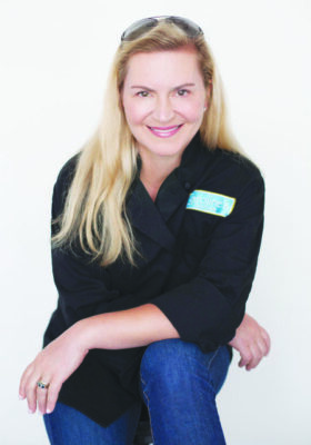 white woman with long blond hair, wearing black chef's coat, sitting in front of gray background, leaning forward smiling