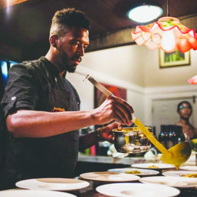 black man standing at counter, ladling sauce onto rows of plates in restaraunt