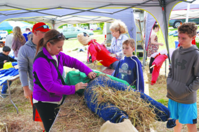 family of man, woman and two boys putting straw into a shirt and pants to make a scarecrow, under tent at community event