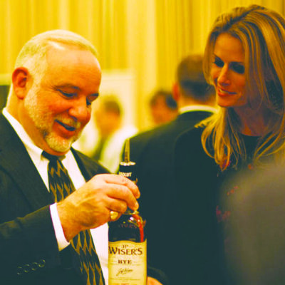 older man holding up liquor bottle as he talks with woman, both in formal wear