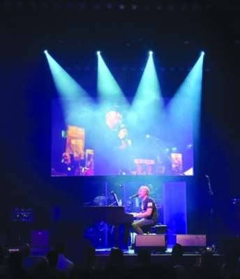 man playing piano on stage with screen behind him showing another man singing into microphone