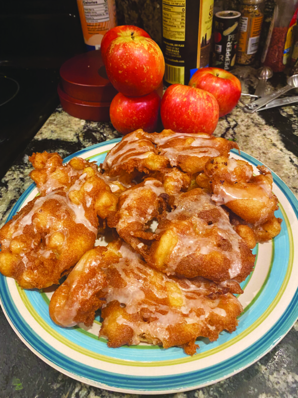 plate piled with apple fritters with drizzled icing, sitting on counter beside apples