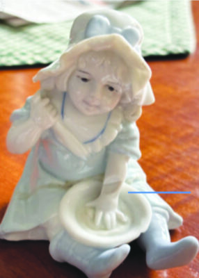 ceramic figurine of young girl wearing dress and bonnet, sitting with legs outstretched, holding plate on her lap and spoon in her other hand