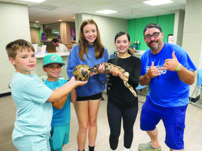 two girls and two boys standing in school hallway holding a long snake between them, adult man posing beside them with his thumbs up