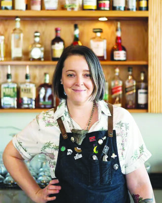 women with shoulder length hair standing in front of shelves with liquor bottles on them, wearing apron with lots of enamel pins on it