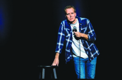 man on dark stage, leaning against stool, holding microphone