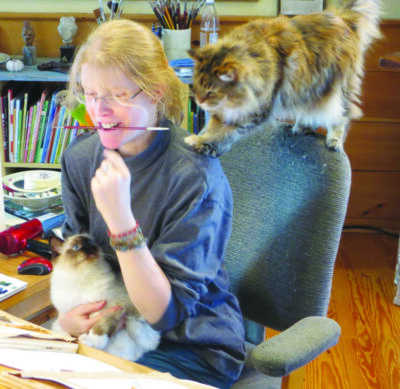 young woman sitting on computer chair with paintbrush held in her mouth, one cat in her lap, another cat standing on the back of the chair, at desk