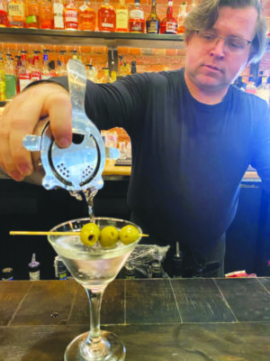 man in black shirt behind restaurant bar pouring ingredients into martini glass with three olives in toothpick along the top