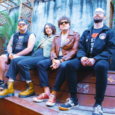 four band members, only one of them a woman, sitting against outdoor wall