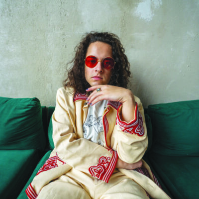 man with long curly hair, wearing red circular glasses and lounge suit, sitting on couch, cement wall behind