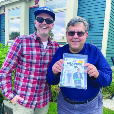 two men standing outside house, one man holding custom made action figure in packaging