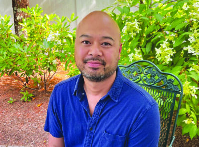 headshot of youngish man, fashionably bald with beard, sitting on chair outside in front of bush