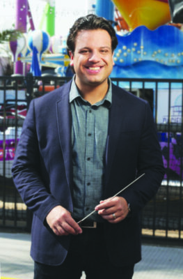 man wearing button up shirt and suit jacket, standing for photo, smiling, holding conductor's baton