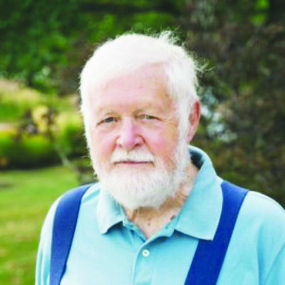 headshot of man with white hair and white beard standing outside in field, blue button up shirt and blue suspenders