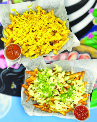 two cardboard plates of french fries, each covered in different sauce topping, with plastic cups of ketchup