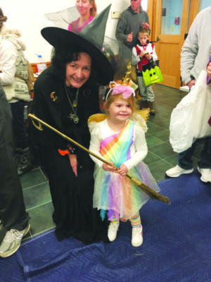 little girl dressed in white floofy costume dress with rainbow stripes across front, headband with unicorn ears and horn, sposing with woman dressed in black with pointy witch hat 