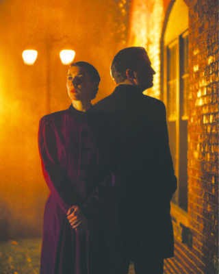 woman and man standing back to back in old fashioned costuming, outside near brick building, lampost giving off warm light behind them, fog making spooky atmosphere