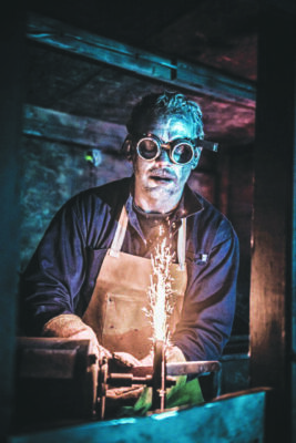 man dressed as ghoulish welder, holding metal rod to grinder to make sparks at haunted house attraction