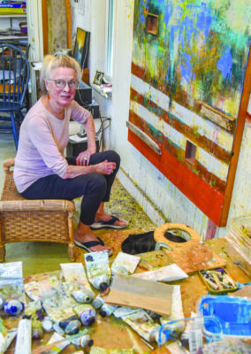 woman sitting on low stool in art studio covered in paint, canvas hanging on wall as she works