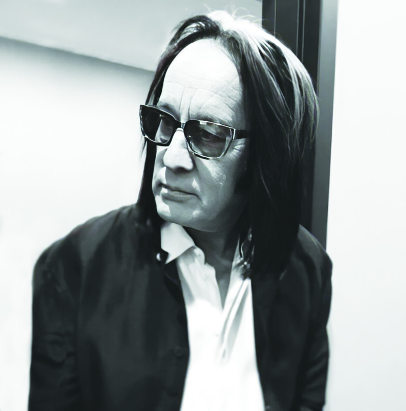 black and white headshot of man with shoulder length hair, wearing sunglasses indoors