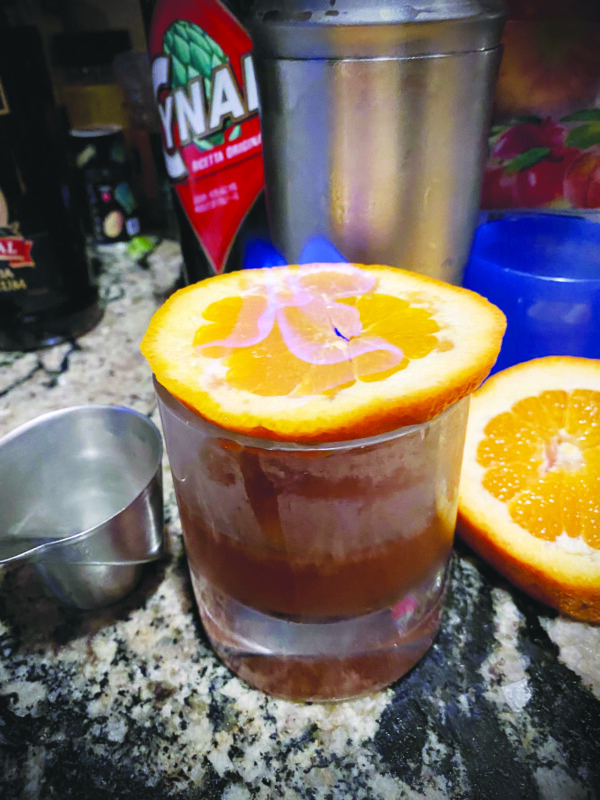 short glass filled with dark colored cocktail, orange slice sitting on rim of glass, blue flames rising from center of orange