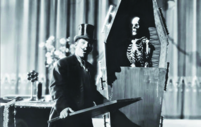 scene from old movie showing man in suit and top hat opening standing coffin with skeleton inside