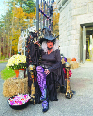 woman wearing orange facepaint, dressed in black witch dress, striped purple and black stockings, and pointy hat, sitting in front of stone tower, bowl of candy beside her, one of her hands raised in spooky beckoning