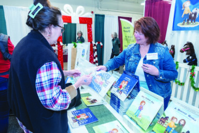 women behind table stacked with children's books, handing bag to female customer at event