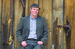 man wearing button down shirt and jacket leaning against barn door and smiling at camera