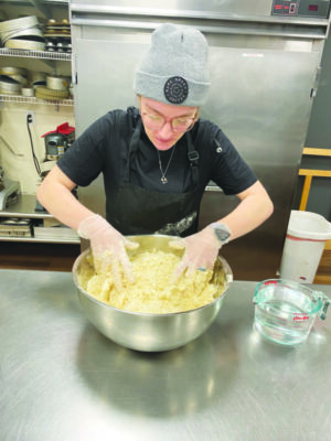 woman wearing knitted hat, standing in industrial kitchen, kneading dough in large stainless steel bowl