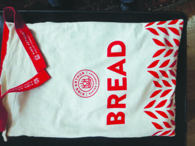 canvas bag with wheat design on bottom and word Bread across the front, laying flat on table, ties at top for closing.