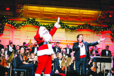 orchestra on stage, conductor at front, man dressed as Santa Claus waving to audience