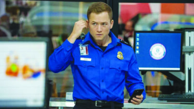 A TSA worker wearing a blue jacket adjusts a microphone in his ear with a serious expression