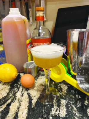 Cocktail in stemmed glass on counter surrounded by lemon, bottle of apple cider, an egg, cocktail shaker