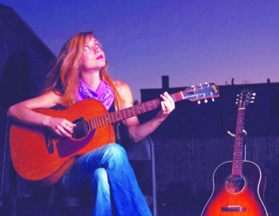 long haired woman sitting in chair in dimly lit room holding guitar on lap, looking upwards while playing