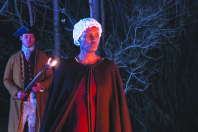 film still of black woman wearing colonial dress and cloak, standing in woodes at night, man in colonial dress standing behind her with light torch