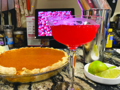 A cranberry margarita in a clear glass sits next to a pumpkin pie and a bowl of limes