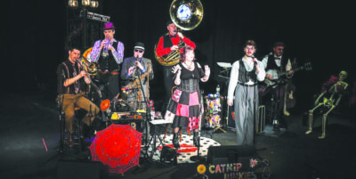 A cat-themed band wearing red and black plays a variety of instruments underneath a spotlight