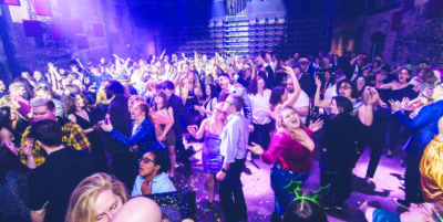 A large crowd of fancily dressed people dances in a purple lit room as confetti floats around