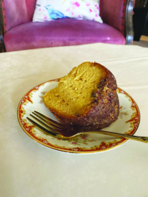 slice of light colored cake with brown topping on decorated china plate, with fork, on table covered with white table cloth