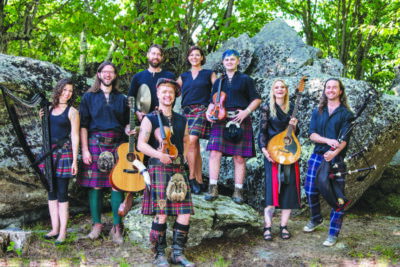 group of people dressed in scottish style garb, standing in front of large rocks outside, holding instruments