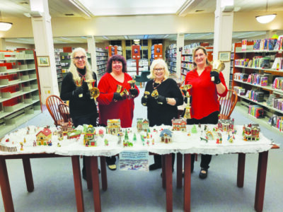 four women, two wearing black shirts and two wearing red shirts, standing in library behind table decorated with ceramic Christmas village, holding ringing bell instruments.