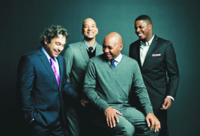 four men wearing suits and ties laughing, dark blue backdrop, studio photo