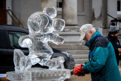 A man in a blue coat uses a saw to carve an ice sculpture of Mickey Mouse