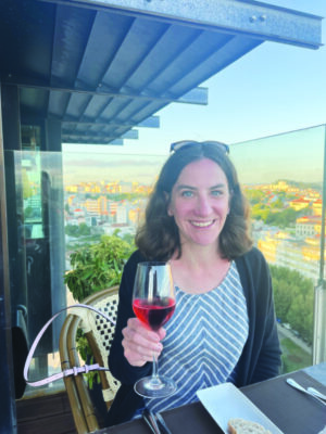 woman sitting at table on balcony overviewing city. She holds up glass of wine, smiling