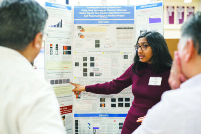 A girl explains her science fair project to two scientists in white coats.