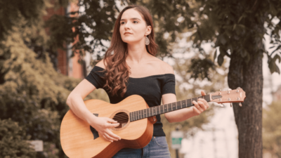 Rebecca Turmel, wearing a shoulder less T, plays the guitar while looking wistfully into the distance