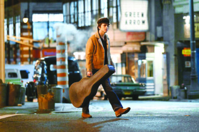 A young man walks across a city parking lot carrying a brown guitar case